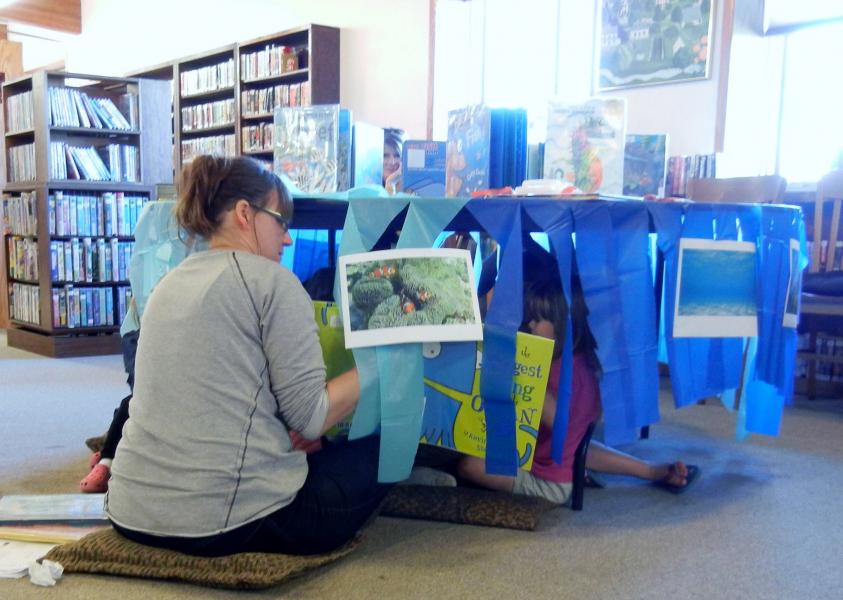 Kids are read stories while sitting under a table with blue streamers to simulate the being in the deepest part of the ocean
